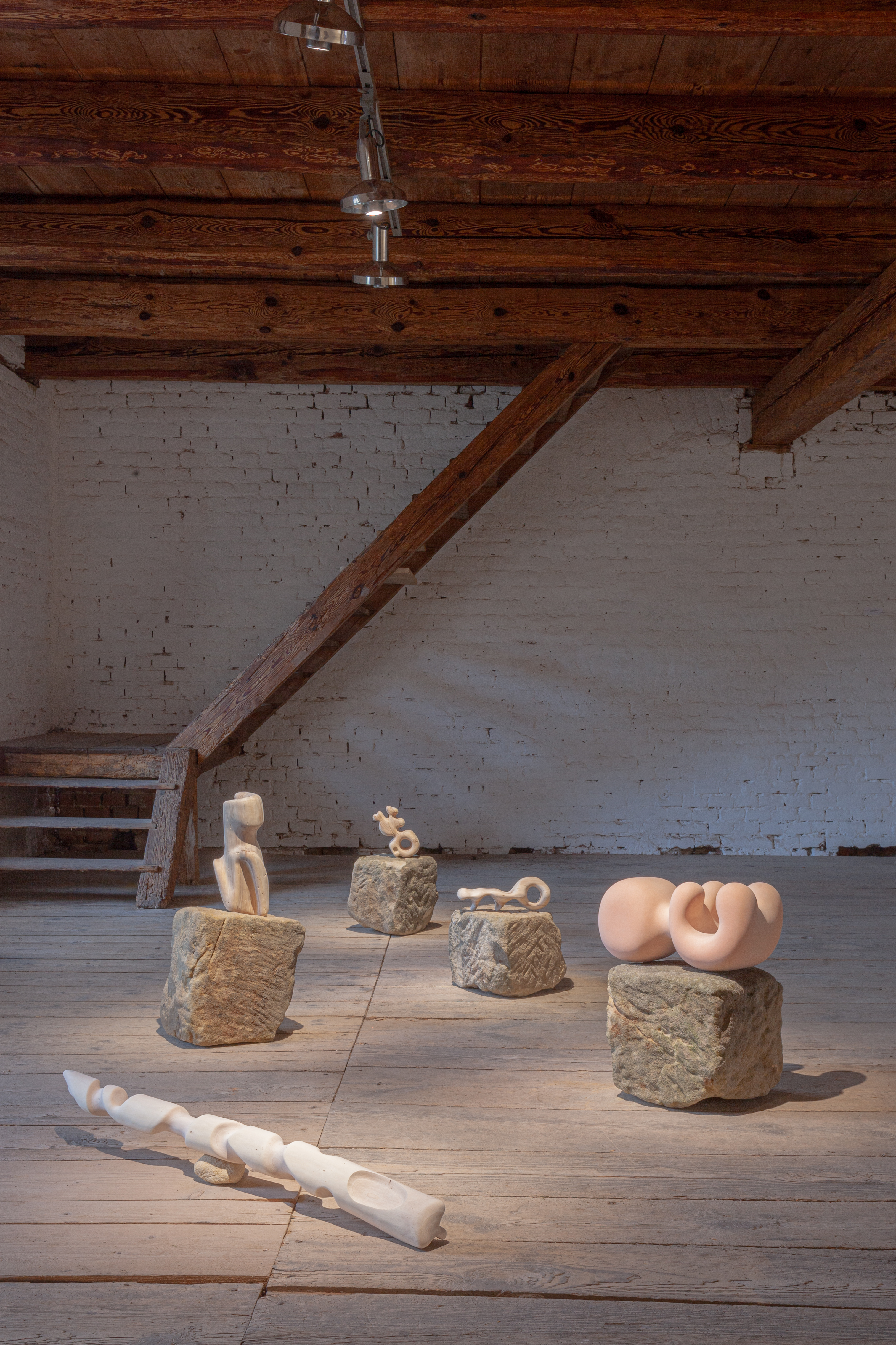 Photo of an art installation called Hands (and Other Products of Labor) 2.0 by Klára Čermáková (Farmstudio Open, Bohemia Farmstudio, Vysoká, Czechia) [Front view]. Five middle-size sculptures/objects of organic shapes (polyurethane foam, willow wood) lie on smaller sandstone pedestals in harmonic composition inside the gallery (originally a medieval barn).