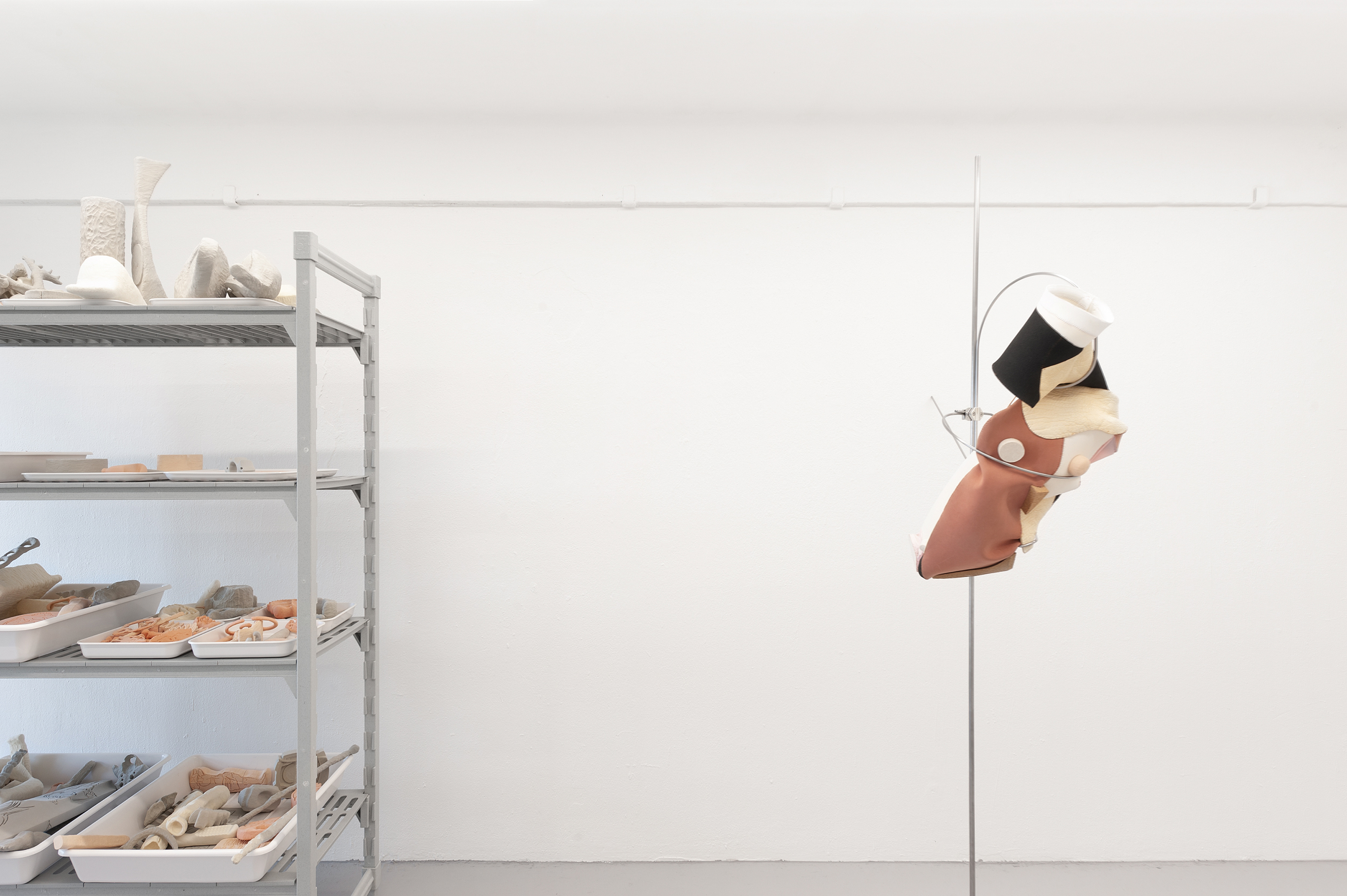 Photo of an art installation called Hands (and Other Products of Labor) by Klára Čermáková (Diploma Projects 2020, UMPRUM — Academy of Arts, Architecture & Design in Prague, Czechia) [Side view]. On the left side, you can see a large laboratory shelf on which many smaller objects depict parts of the human body, bones, objects of daily use, or their free fusion. On the right side is a sculpture of the torso (fragment of the human body) on a metal base.