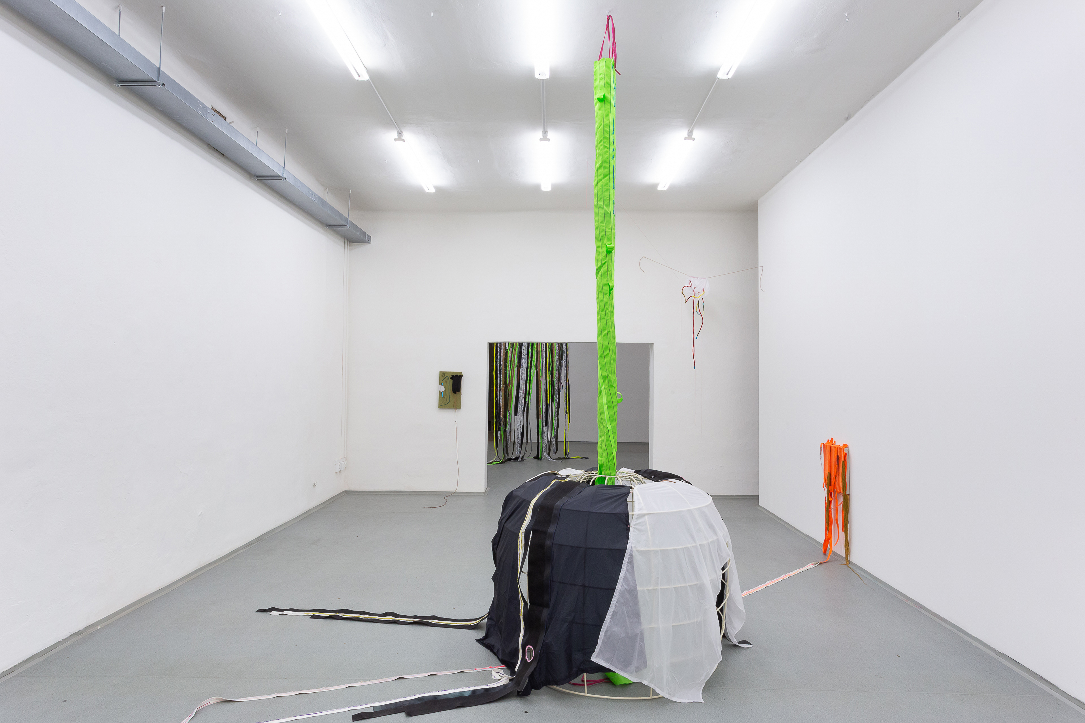 Photo of an art installation called Living Tent by Klára Čermáková (Galerie FaVu, Brno, Czechia, 2018) [Back view]. The central object in the photo is a metal skeleton covered with fabric (a kind of tent or shelter) sewn together from many smaller pieces of fabric/material. This cover then freely continues/spreads to different sides of the room, up to some stitched paintings (hung on the walls) or ceiling.